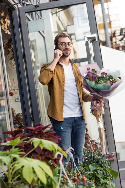 Souriant bel homme sortir de magasin de fleurs avec bouquet et parler par smartphone — Photo de stock