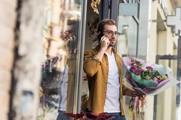 Hombre guapo saliendo de la florería con ramo y hablando por teléfono inteligente - foto de stock