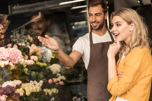 Florista feliz apontando flores para o cliente animado na vitrine na loja de flores — Fotografia de Stock