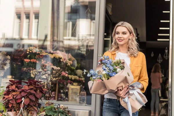 Bela mulher saindo da loja de flores com buquê embrulhado e olhando para longe — Fotografia de Stock