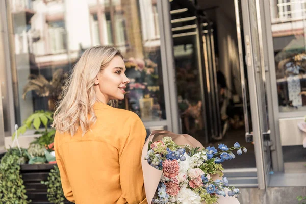 Vista trasera de chica atractiva caminando con ramo envuelto cerca de floristería - foto de stock