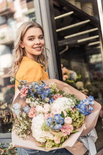 Chica atractiva sosteniendo hermoso ramo cerca de la tienda de flores y mirando a la cámara - foto de stock
