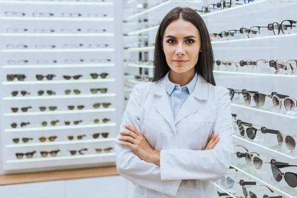 Professioneller Augenoptiker mit verschränkten Armen, der in der Nähe von Regalen mit Sehkraft in Optik posiert — Stockfoto