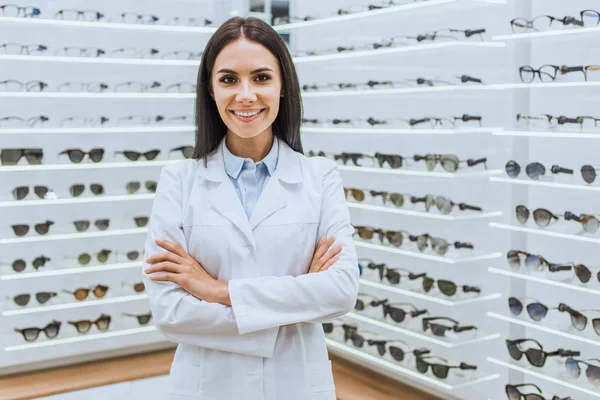 Profissional optometrista sorridente posando com braços cruzados perto de prateleiras com visão na óptica — Fotografia de Stock