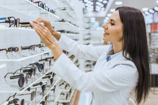 Hermosa óptica sonriente tomando gafas de los estantes en la tienda oftálmica - foto de stock