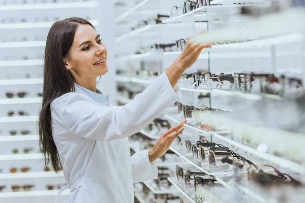 Professional smiling optician taking glasses from shelves in ophthalmology — Stock Photo