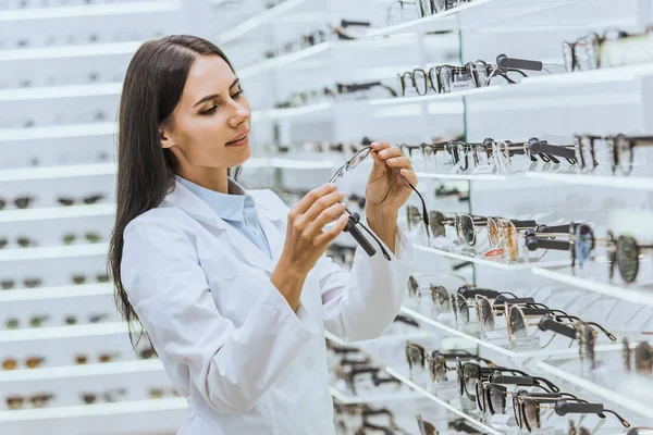 Beautiful optometrist looking at eyeglasses near shelves in ophthalmic shop — Stock Photo