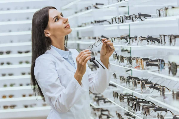 Schöne professionelle Arzt nimmt Brillen aus Regalen in Optik — Stockfoto