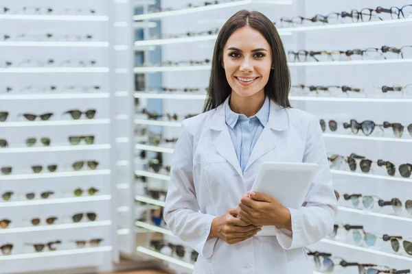 Attraktiver professioneller Optiker mit digitalem Tablet vor der Kamera im Augenoptikergeschäft — Stockfoto