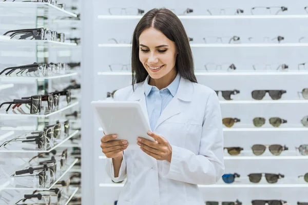 Beautiful female optometrist using digital tablet near shelves with eyesight in ophthalmic shop — Stock Photo