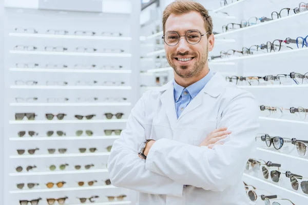 Portrait d'optométriste professionnel souriant dans des lunettes posant avec les bras croisés dans l'optica — Photo de stock