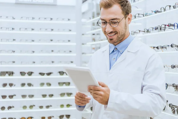 Smiling male optician using digital tablet in optica with eyeglasses on shelves — Stock Photo