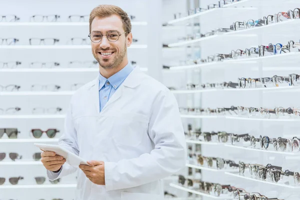 Optometrista masculino sorridente usando comprimido em óptica com visão nas prateleiras — Fotografia de Stock