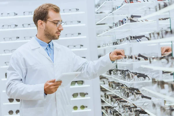 Professional optician with tablet taking glasses from shelves in optica — Stock Photo