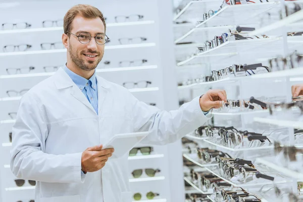 Professioneller Augenoptiker mit digitalem Tablet, der Brillen aus den Regalen in der Optik nimmt — Stockfoto