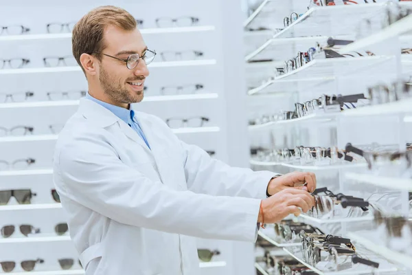 Oculista sorridente che prende la vista da ripiani in negozio oftalmico — Foto stock