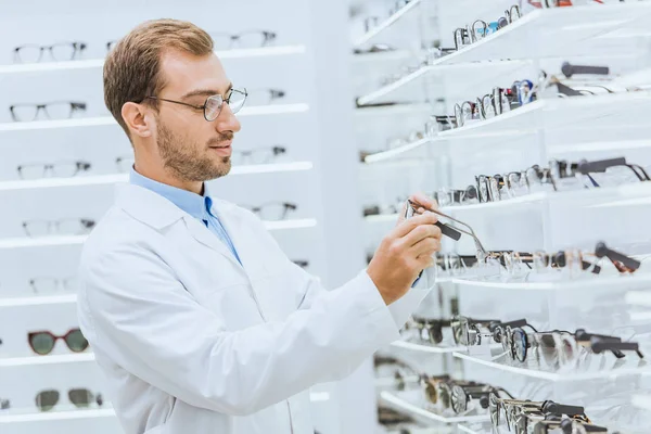 Professional male oculist taking eyeglasses from shelves in ophthalmic shop — Stock Photo