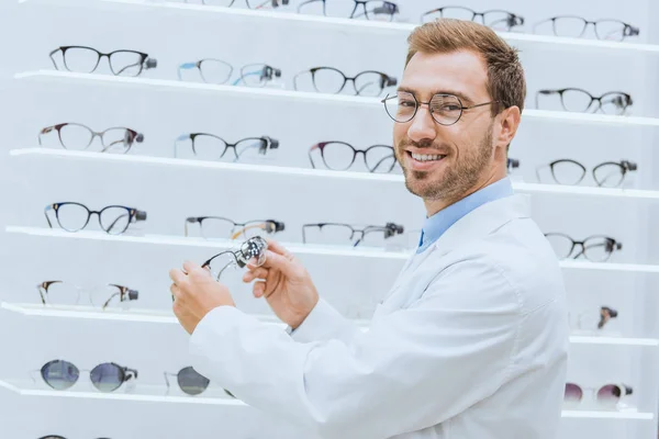 Médico sonriente en bata blanca tomando anteojos de estantes en óptica - foto de stock