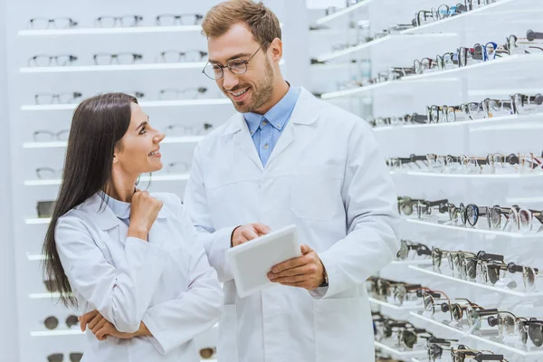 Dos optometristas usando tableta digital y mirándose entre sí en óptica con gafas - foto de stock