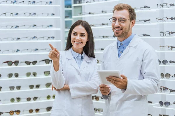 Optometristas sorridentes usando tablet digital, enquanto o médico feminino apontando para óculos na óptica — Fotografia de Stock