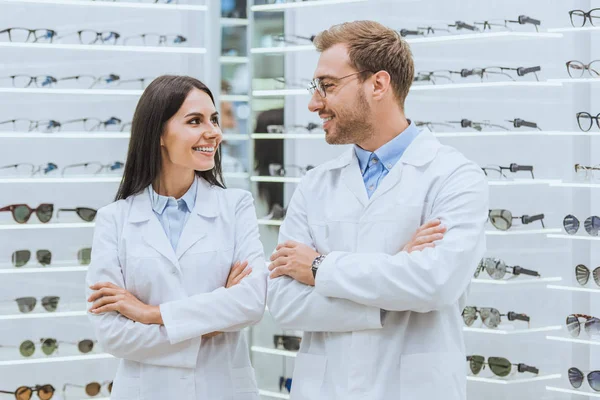 Professional smiling doctors posing with crossed arms in ophthalmic shop with eyeglasses — Stock Photo