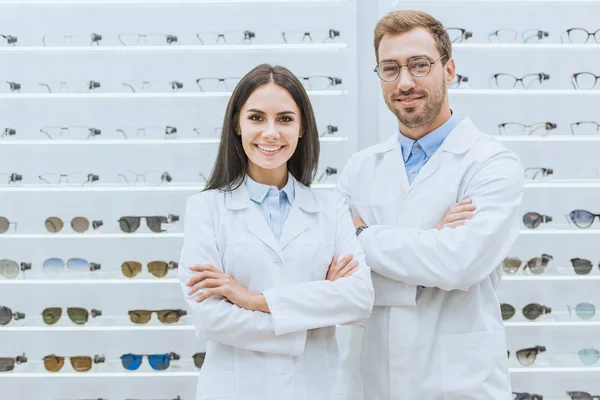 Professional smiling oculists posing with crossed arms in ophthalmic shop with glasses — Stock Photo