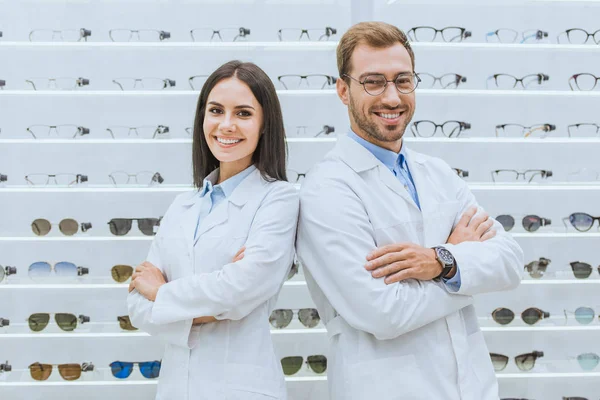 Professional opticians posing with crossed arms in ophthalmic shop with glasses — Stock Photo