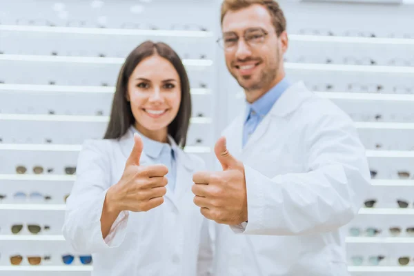 Ópticos profesionales mostrando pulgares hacia arriba en la tienda oftálmica con vista en los estantes - foto de stock