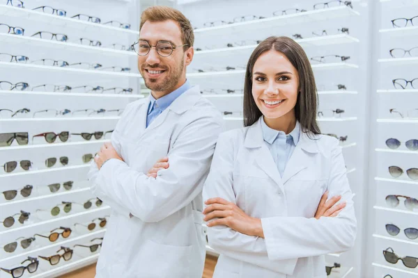 Ópticos profesionales en batas blancas de pie con brazos cruzados en oftalmología - foto de stock