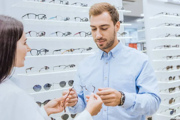 Partial view of female oculist giving eyeglasses to young man in optica — Stock Photo