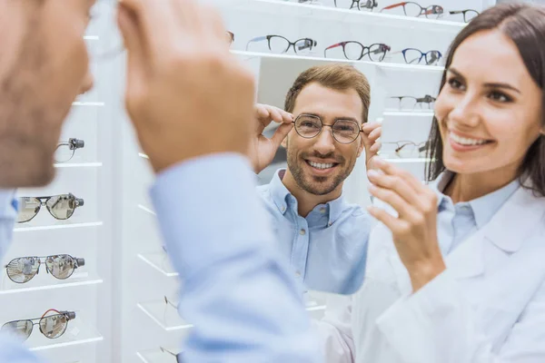 Весёлая женщина-оптометрист держит зеркало, улыбаясь мужчине, выбирающему очки в офтальмологическом магазине — стоковое фото