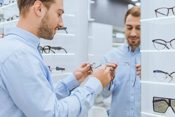 Hombre sonriente eligiendo anteojos delante del espejo en la tienda oftálmica - foto de stock