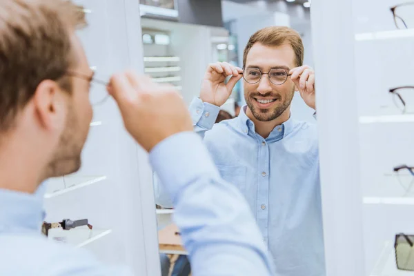 Partial view of smiling young man choosing eyeglasses and looking at mirror in optica — Stock Photo