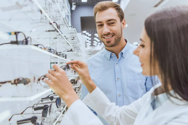 Side view of female oculist taking eyeglasses from shelves to happy man in ophthalmic shop — Stock Photo