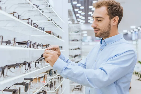 Вибірковий фокус молодого щасливого чоловіка, який бере окуляри з полиць в офтальмологічному магазині — стокове фото