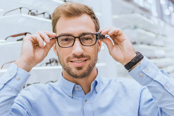 Portrait of young man choosing eyeglasses and looking at camera in optics — Stock Photo