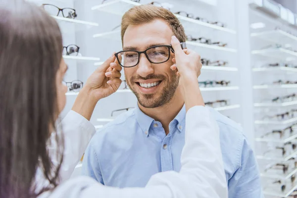 Partial view of female optometrist putting on eyeglasses on smiling man in optics — Stock Photo