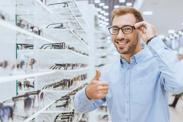 Happy young man choosing eyeglasses and doing thumb up gesture in optica — Stock Photo