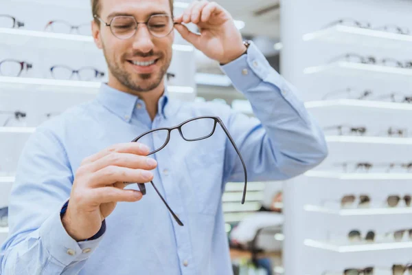 Selective focus of handsome young man choosing eyeglasses in ophthalmic shop — Stock Photo
