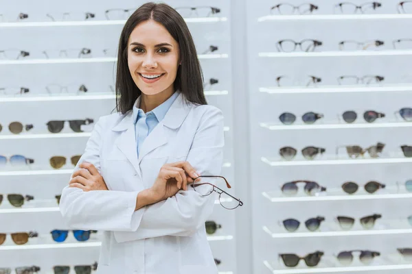 Óptico sonriente profesional sosteniendo gafas cerca de estantes en óptica - foto de stock