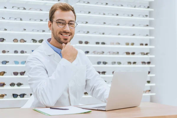 Opticien professionnel utilisant un ordinateur portable à la table avec des papiers dans la boutique ophtalmique — Photo de stock