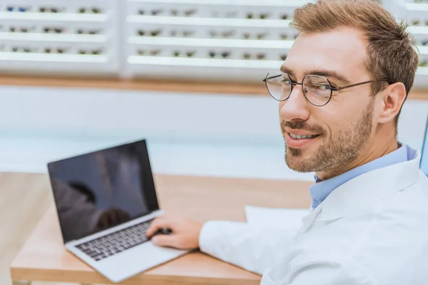 Optometrista guapo profesional usando el ordenador portátil en la tienda oftálmica - foto de stock