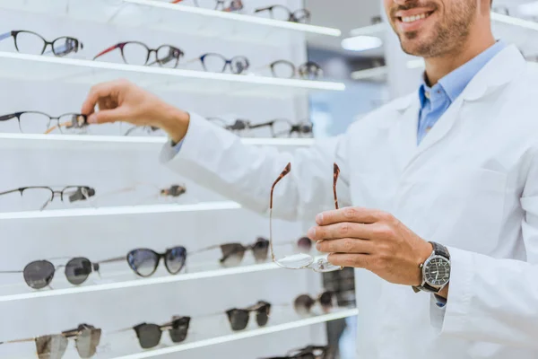 Vista recortada del optometrista tomando anteojos de estantes en la tienda oftálmica - foto de stock