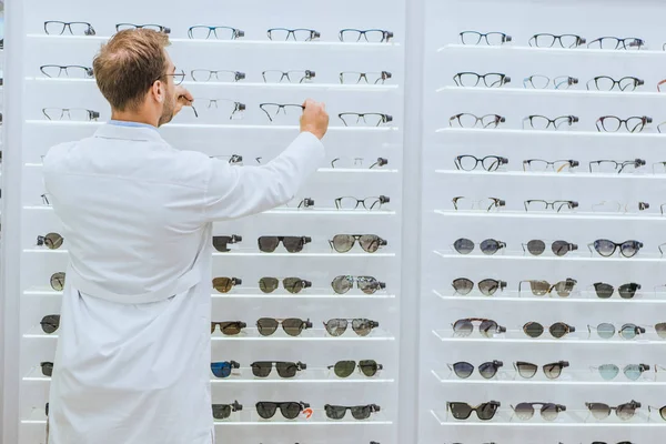 Back view of oculist in white coat taking glasses from shelves in ophthalmic shop — Stock Photo