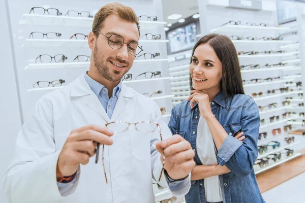 Hermoso macho oculista mostrando gafas a joven mujer en oftalmología tienda - foto de stock