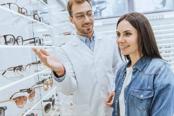 Male oculist pointing by hand at shelves with eyeglasses to female in optics — Stock Photo