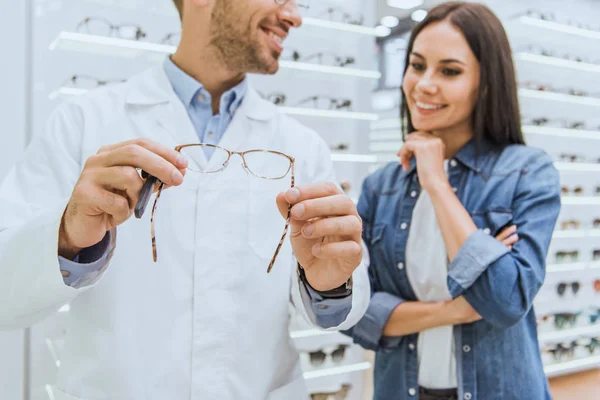 Partial view of happy male oculist helping female to choose pair of eyeglasses in ophthalmic shop — Stock Photo