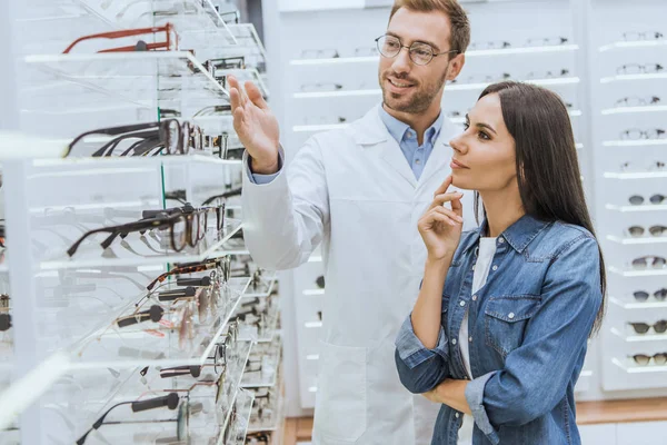 Young male optometrist pointing by hand at shelves with eyeglasses to woman in optics — Stock Photo