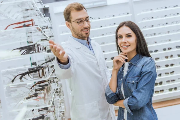 Alegre oculista masculino apuntando a mano a los estantes con gafas a la mujer en óptica - foto de stock