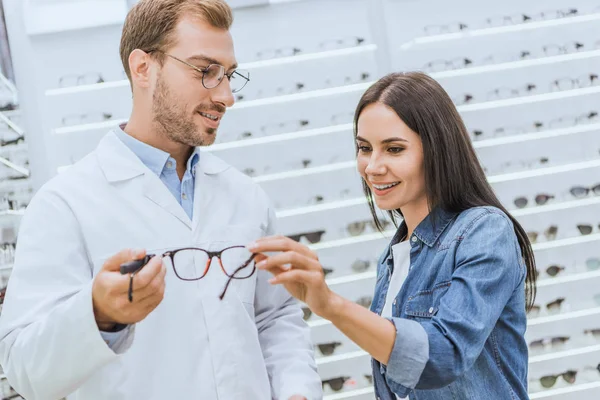 Foco seletivo do optometrista masculino mostrando óculos para mulher sorridente na óptica — Fotografia de Stock